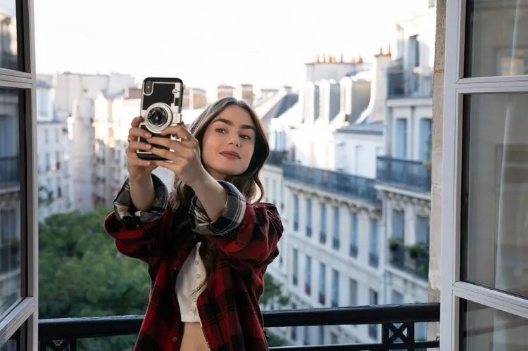 Cena de Emily em Paris, com Emily tirando uma selfie e, ao fundo, prédios históricos da cidade.