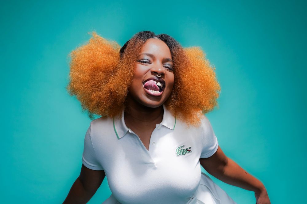 Mulher negra de cabelo ruivo com camiseta gola polo branca sorrindo para a câmera e mostrando a língua