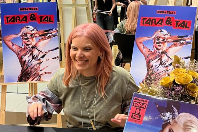 Duda Beat sorrindo entre cópias do disco Tara e Tal em sessão de autógrafos