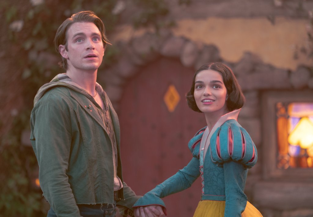 Jonathan (Andrew Burnap) e Branca de Neve (Rachel Zegler) em cena do live-action; eles estão sorrindo de mãos dadas olhando para o lado