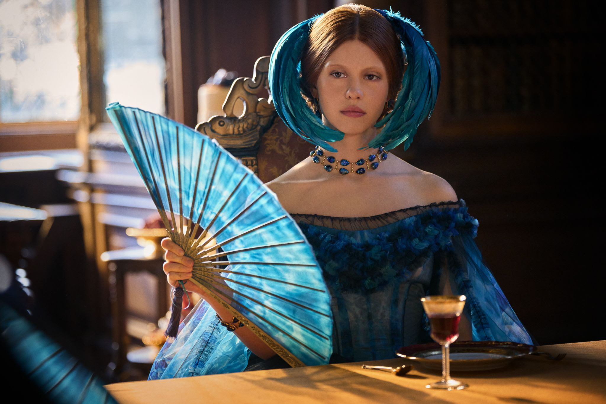 Mia Goth em Frankenstein Mia Goth usa vestido cheio de babados, somados a um laque, um colar e penas azuis na cabeça.