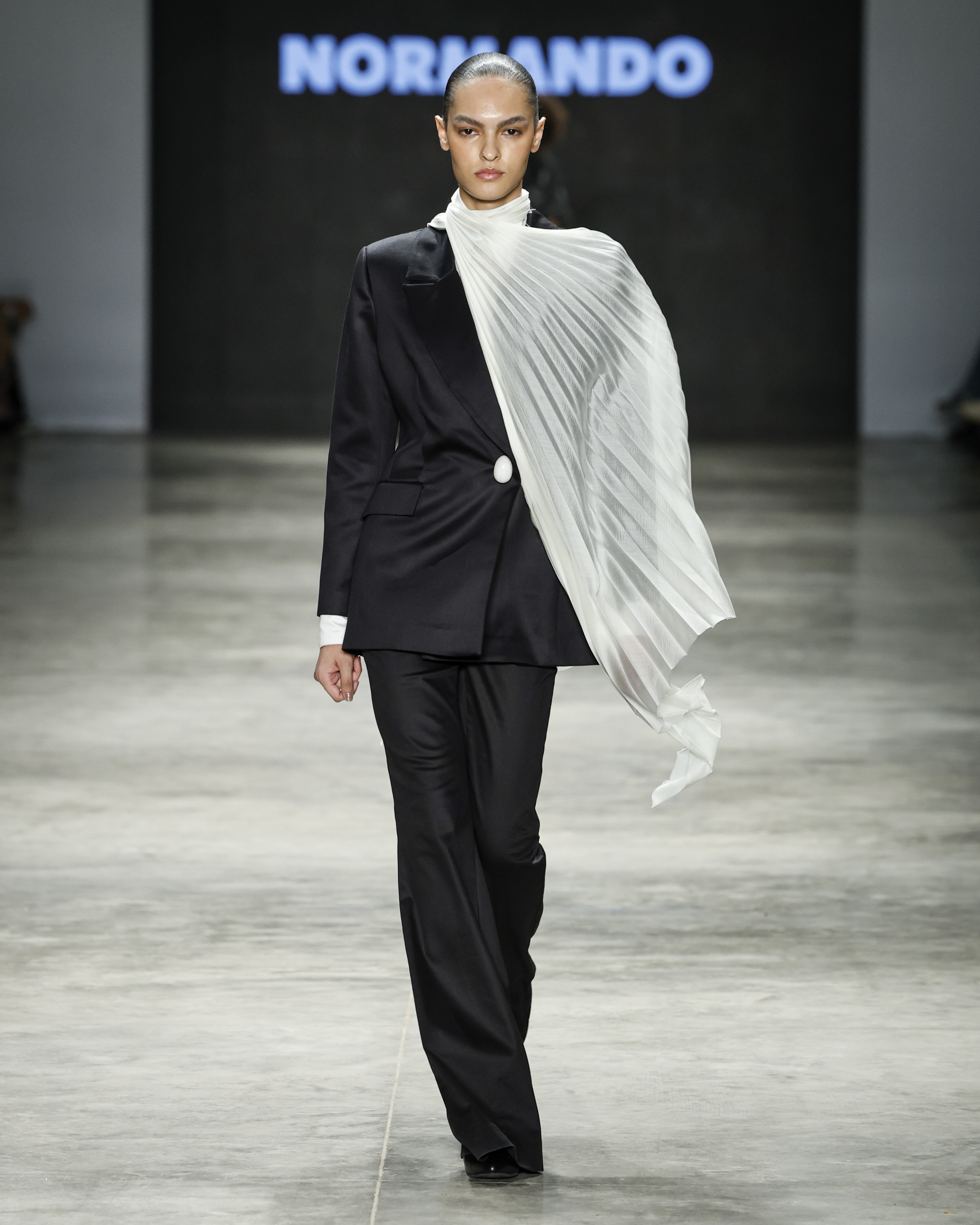 Modelo desfilando na SPFW com look de alfaiataria preto com detalhes brancos 