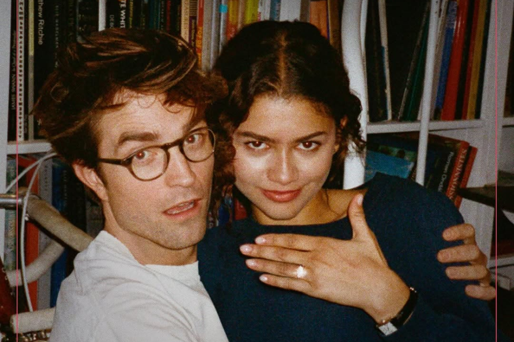 Zendaya e Robert Pattinson estão abraçados, ele usa roupa branca e ela roupa preta, enquanto exibe um anel de noivado