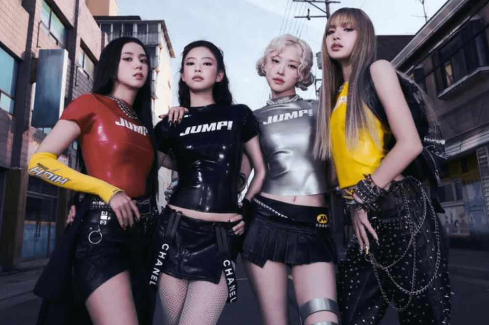 Quatro mulheres posam juntas em uma rua urbana, usando looks modernos e futuristas em cores vermelho, preto, prata e amarelo. Elas vestem tops justos com a palavra “JUMP!” e saias ou shorts com detalhes em couro e correntes, olhando diretamente para a câmera com atitude confiante. Prédios e fios elétricos aparecem ao fundo, reforçando o clima urbano da cena.