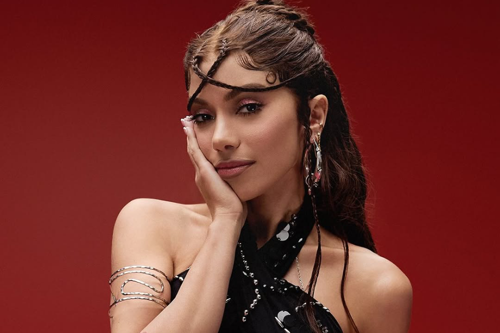 Mulher posa diante de fundo vermelho com a mão apoiada no rosto; ela usa penteado com tranças finas na testa, brincos longos e acessórios prateados no braço, além de look preto.