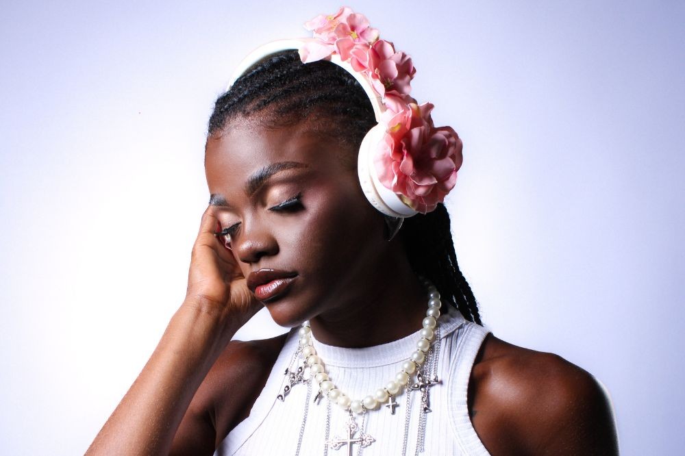 Mulher negra com tranças, olhos fechados e expressão serena, usando fones de ouvido brancos decorados com flores cor-de-rosa, colar de pérolas e pingentes de cruz, em fundo branco