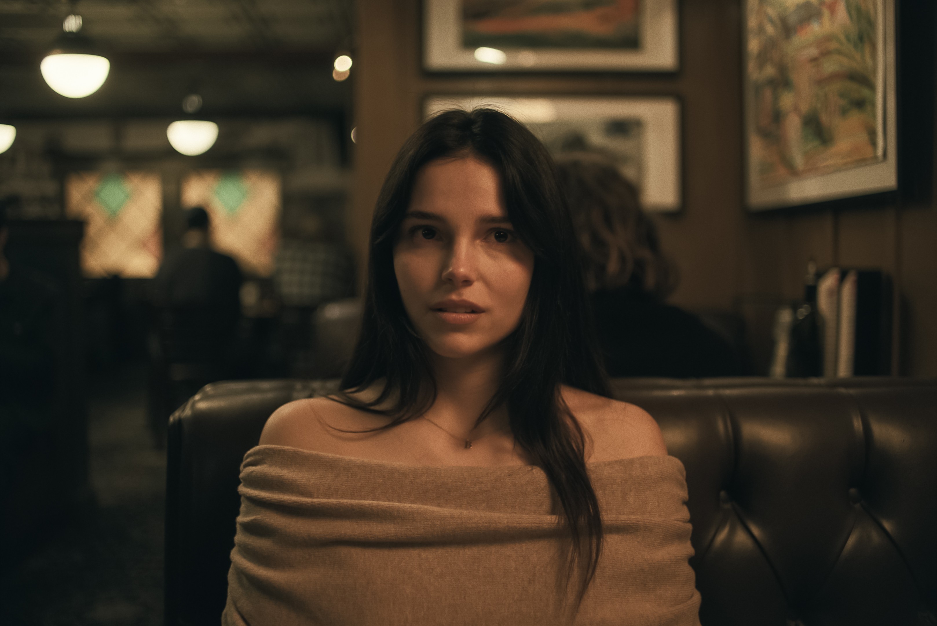 Mulher jovem de cabelos escuros e longos, com blusa bege de ombros à mostra, sentada em um sofá de couro marrom em um restaurante ou bar com pouca luz. Ela olha diretamente para frente com expressão séria. Ao fundo, luzes penduradas e quadros na parede, com pessoas desfocadas