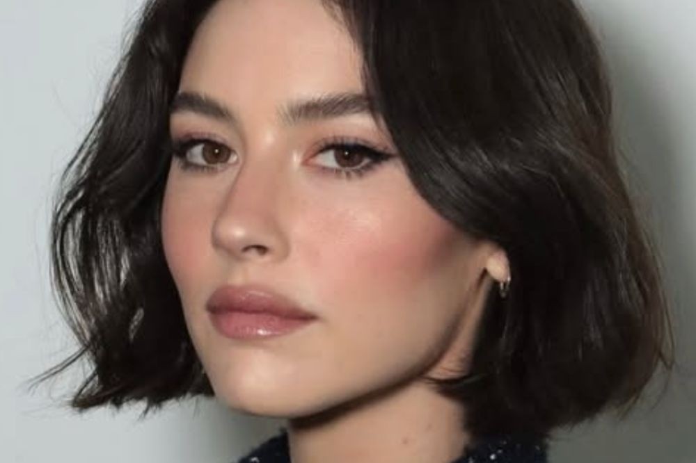 Rosto de uma mulher jovem com cabelo castanho escuro curto e ondulado, maquiagem leve com delineador e batom nude, e um brinco de argola prateado na orelha direita