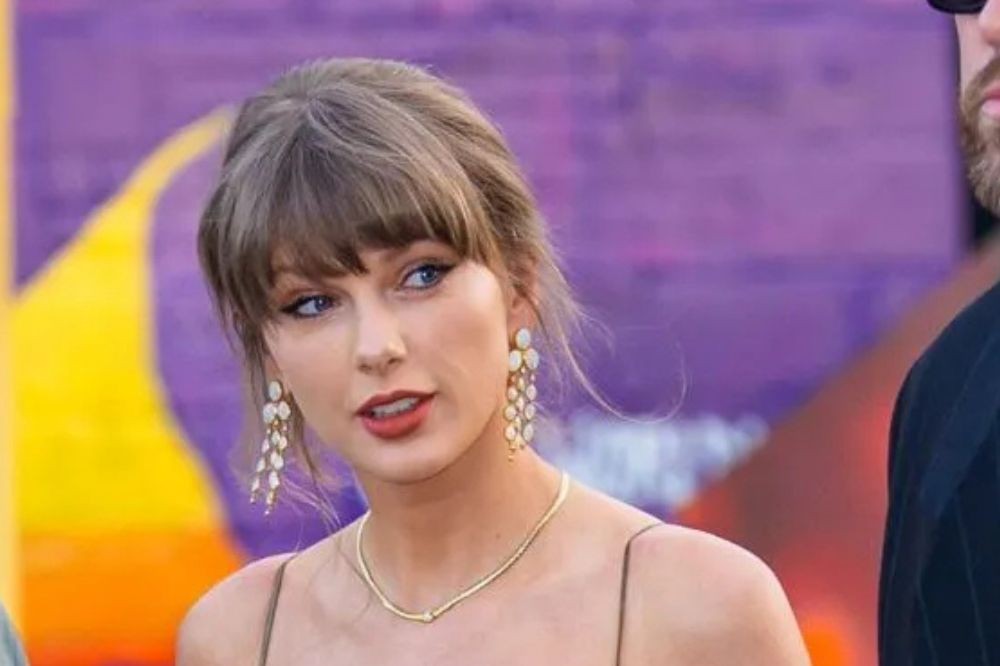 Taylor Swift com cabelo preso e franja, usando brincos longos e colar dourados, maquiagem leve e batom vermelho, olhando para a direita com expressão séria. Ao fundo, um borrão roxo e amarelo