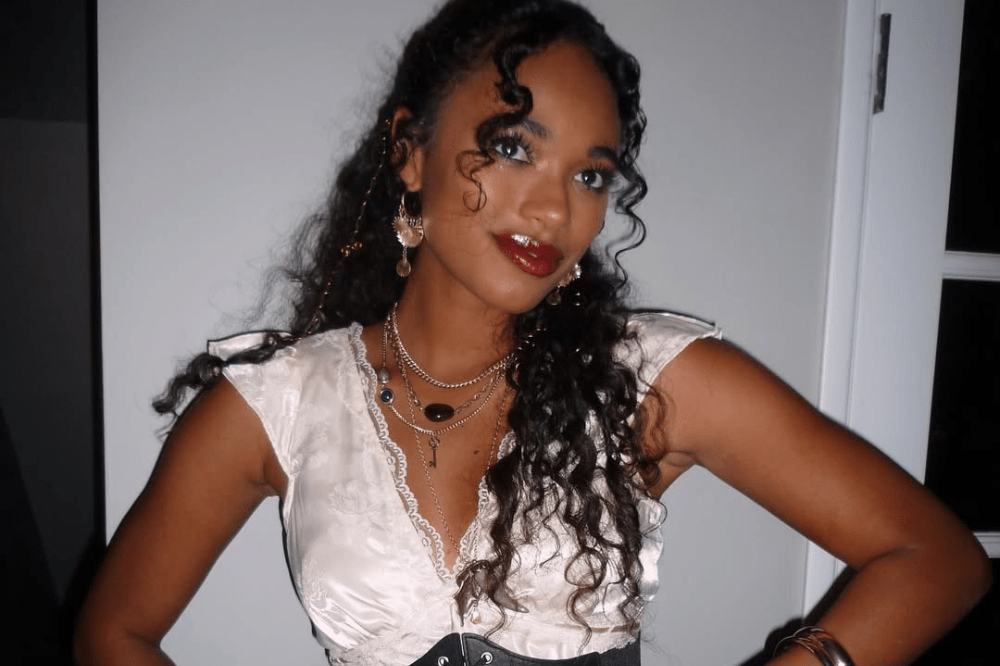 Mulher jovem de pele morena e cabelos longos e cacheados, com maquiagem marcante e batom vermelho, usando blusa de seda clara com renda, vários colares e brincos, posando com as mãos na cintura