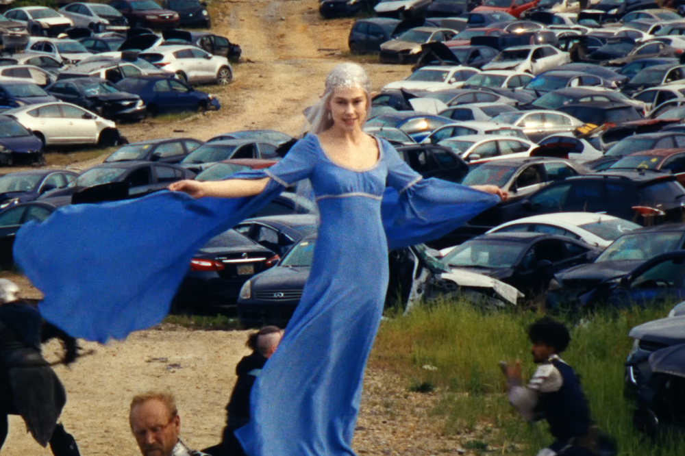 Mulher loira com tiara prateada e vestido azul esvoaçante em pátio de carros batidos, com outras pessoas ao redor