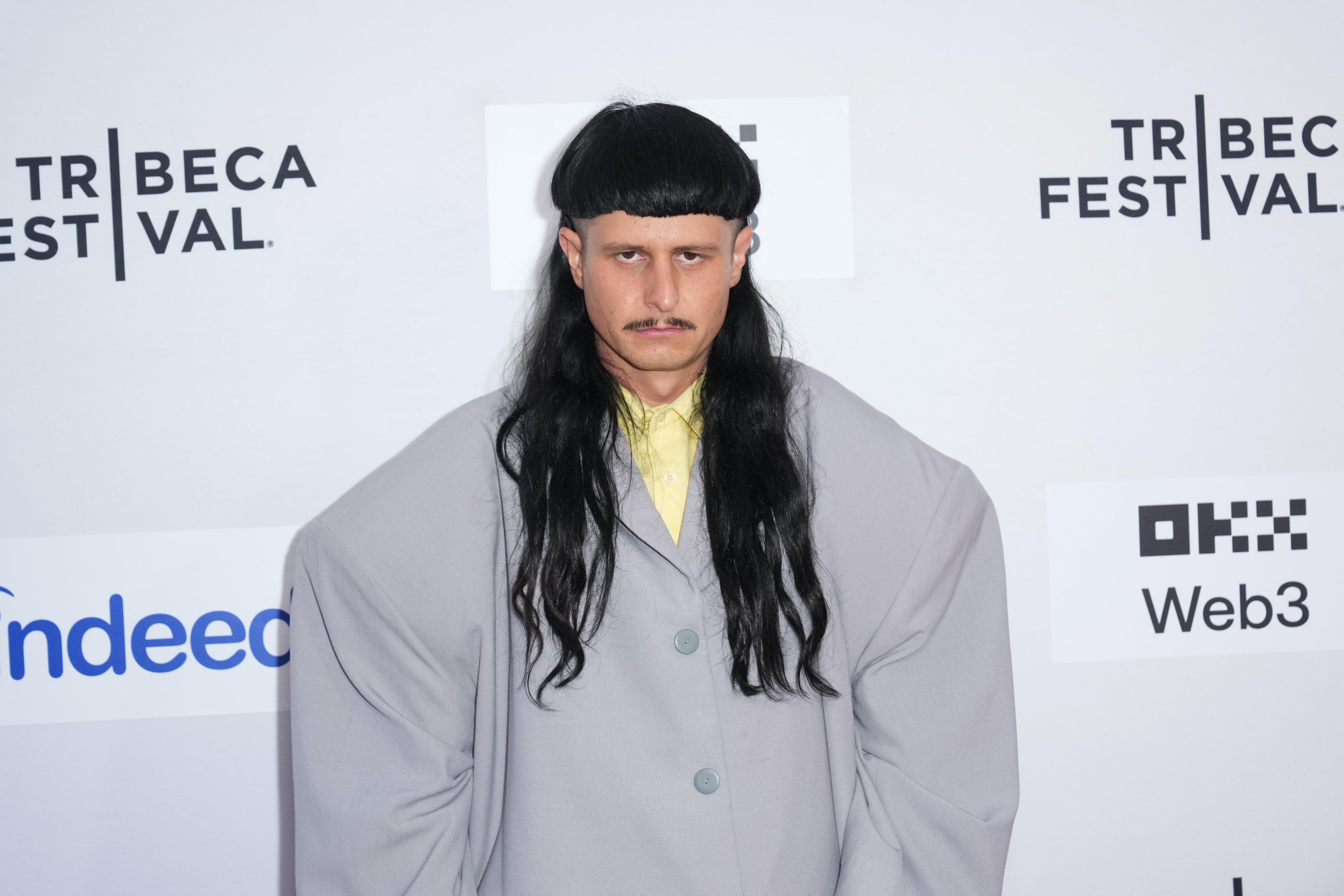 Homem com cabelo preto liso, corte tigela na frente e longo atrás, bigode fino, vestindo camisa amarela e paletó cinza oversized, em frente a um painel branco com logos do Tribeca Festival, Indeed e OKX Web3