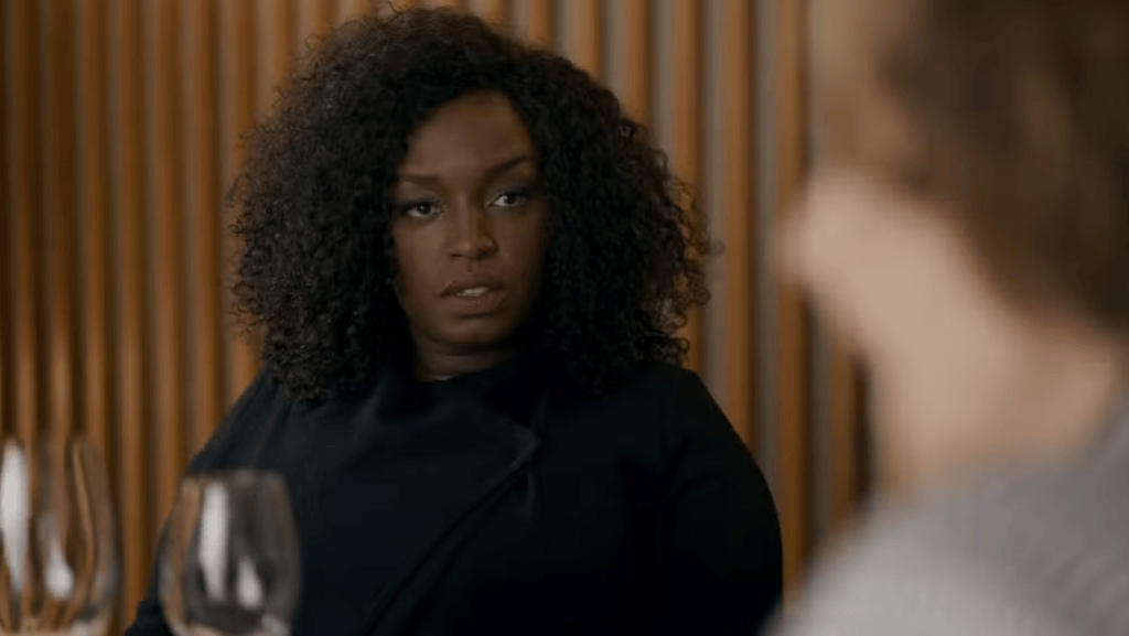 Mulher negra de cabelo cacheado escuro, vestindo casaco preto, olha com expressão séria para alguém fora do enquadramento, com taças de vinho em primeiro plano e parede de madeira ao fundo