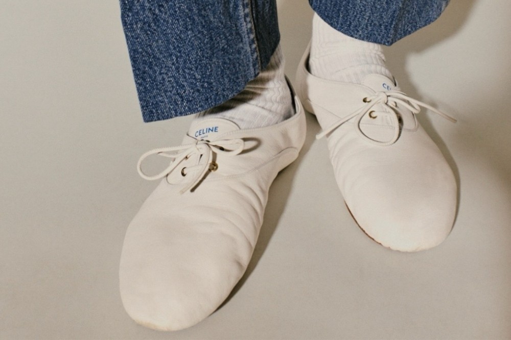 Pés de uma pessoa usando calças jeans azuis, meias brancas e sapatos brancos de couro com cadarços, sobre um fundo cinza claro. O nome CELINE está visível na língua de um dos sapatos