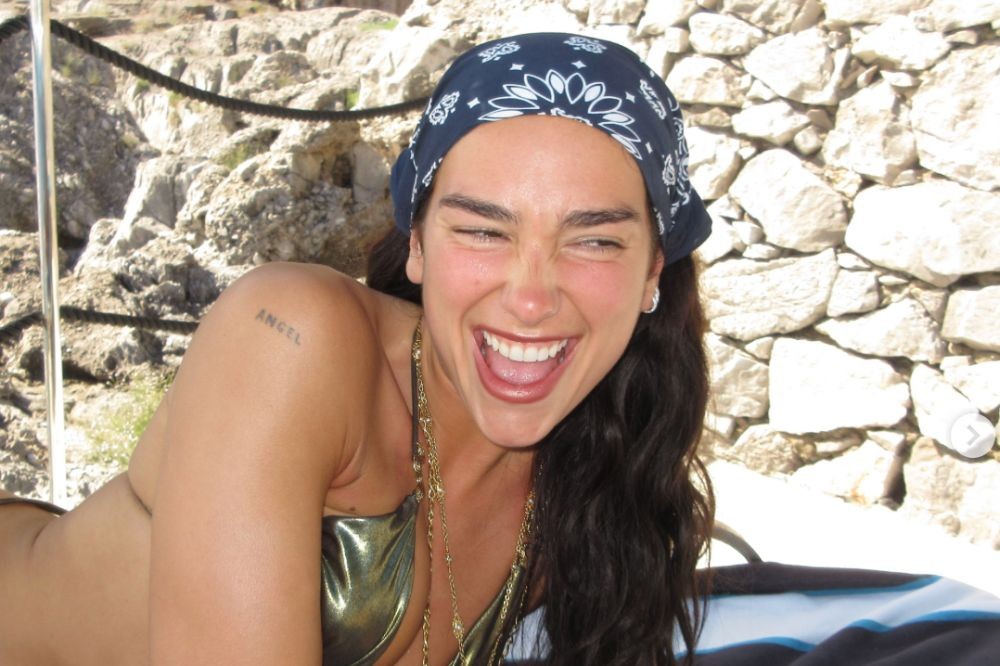 Dua Lipa sorrindo amplamente, com os olhos semicerrados, usando um biquíni dourado, bandana azul escura com estampa branca e um colar dourado. Ela tem uma tatuagem ANGEL no ombro esquerdo e está em um barco, com uma parede de pedras claras ao fundo