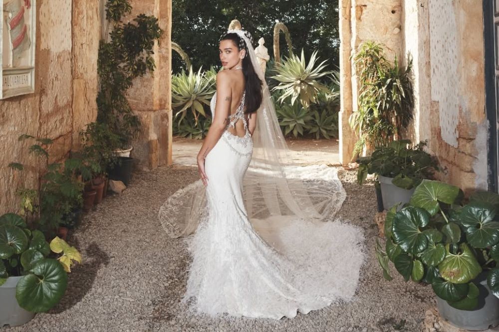 Mulher de cabelos escuros, com um vestido de noiva branco sereia, decote nas costas e véu longo, posa de costas olhando para a câmera em um pátio de pedras com paredes de tijolos e plantas verdes em vasos