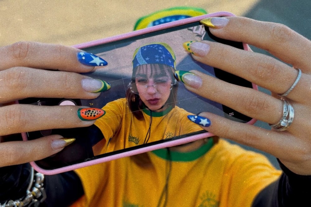 Mãos femininas com unhas decoradas em temas brasileiros seguram um celular rosa, exibindo a tela com uma selfie. A mulher na selfie usa bandana da bandeira do Brasil, óculos e camisa amarela da seleção, com expressão séria. As unhas têm estrelas, melancia e francesinha verde e amarela.