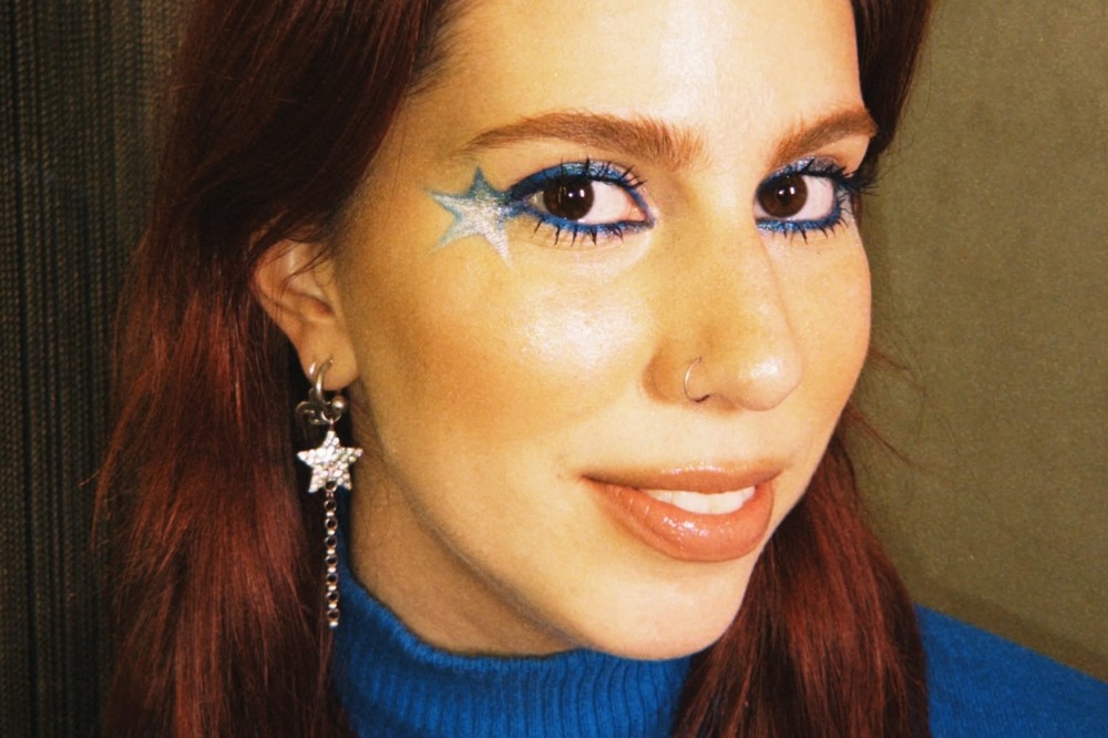 Mulher jovem de cabelos ruivos e pele clara, com maquiagem azul vibrante nos olhos e uma estrela brilhante no canto externo. Ela usa um brinco de estrela prateada com corrente e um suéter azul, sorrindo levemente para a câmera