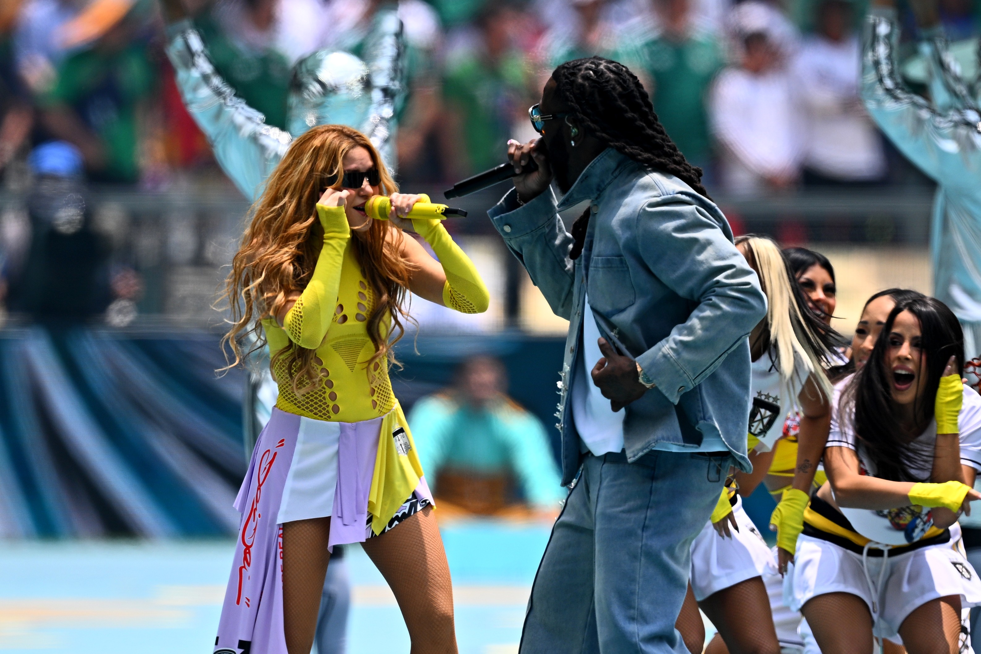 Shakira, de cabelo ondulado e óculos escuros, canta em microfone amarelo, vestindo top neon e saia colorida, ao lado de um homem de dreads e jaqueta jeans, também cantando. Dançarinas e público ao fundo em um estádio