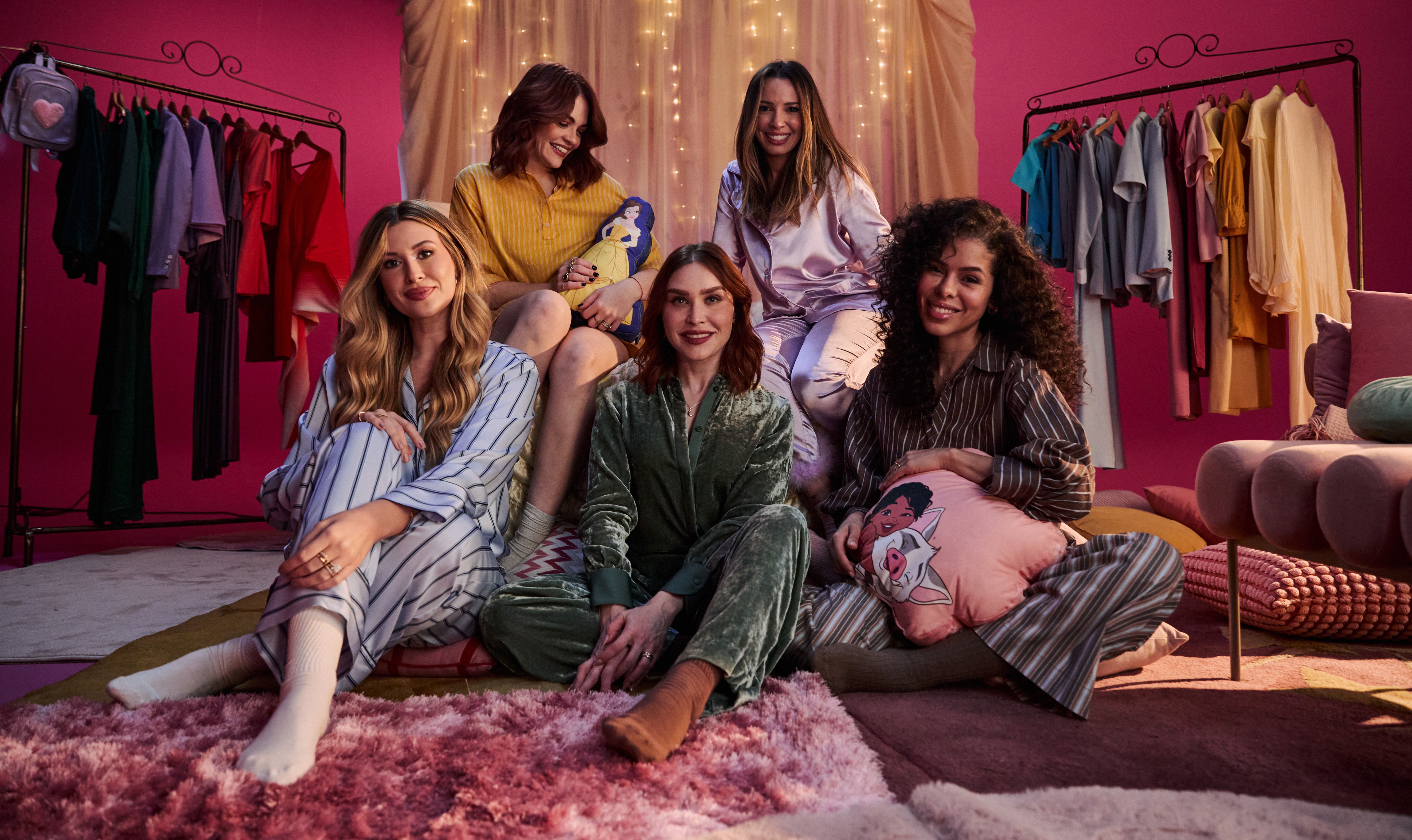 Cinco mulheres sorridentes em pijamas, sentadas em um quarto com fundo rosa e luzes decorativas. Duas mulheres estão sentadas no chão, uma de pijama listrado azul e branco e outra de pijama verde de veludo. Três mulheres estão sentadas atrás delas, uma de pijama amarelo, outra de pijama lilás e a última de pijama listrado marrom e branco. Há um cabideiro com roupas coloridas à esquerda e um sofá rosa à direita. Todas olham para frente, com expressão alegre