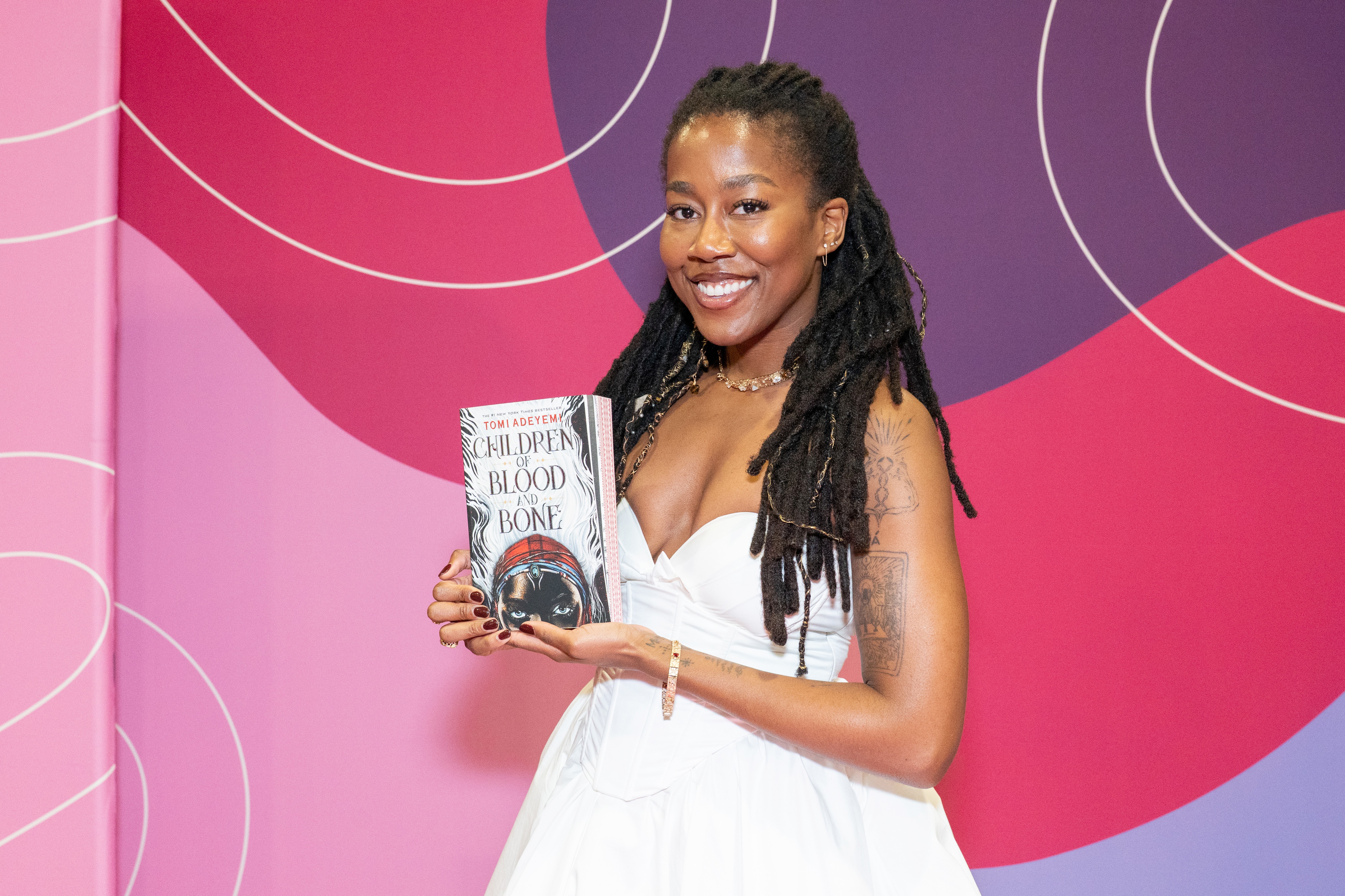 Mulher negra sorridente com dreadlocks, vestindo um vestido branco tomara que caia, segura o livro Children of Blood and Bone contra um fundo rosa e roxo com linhas brancas