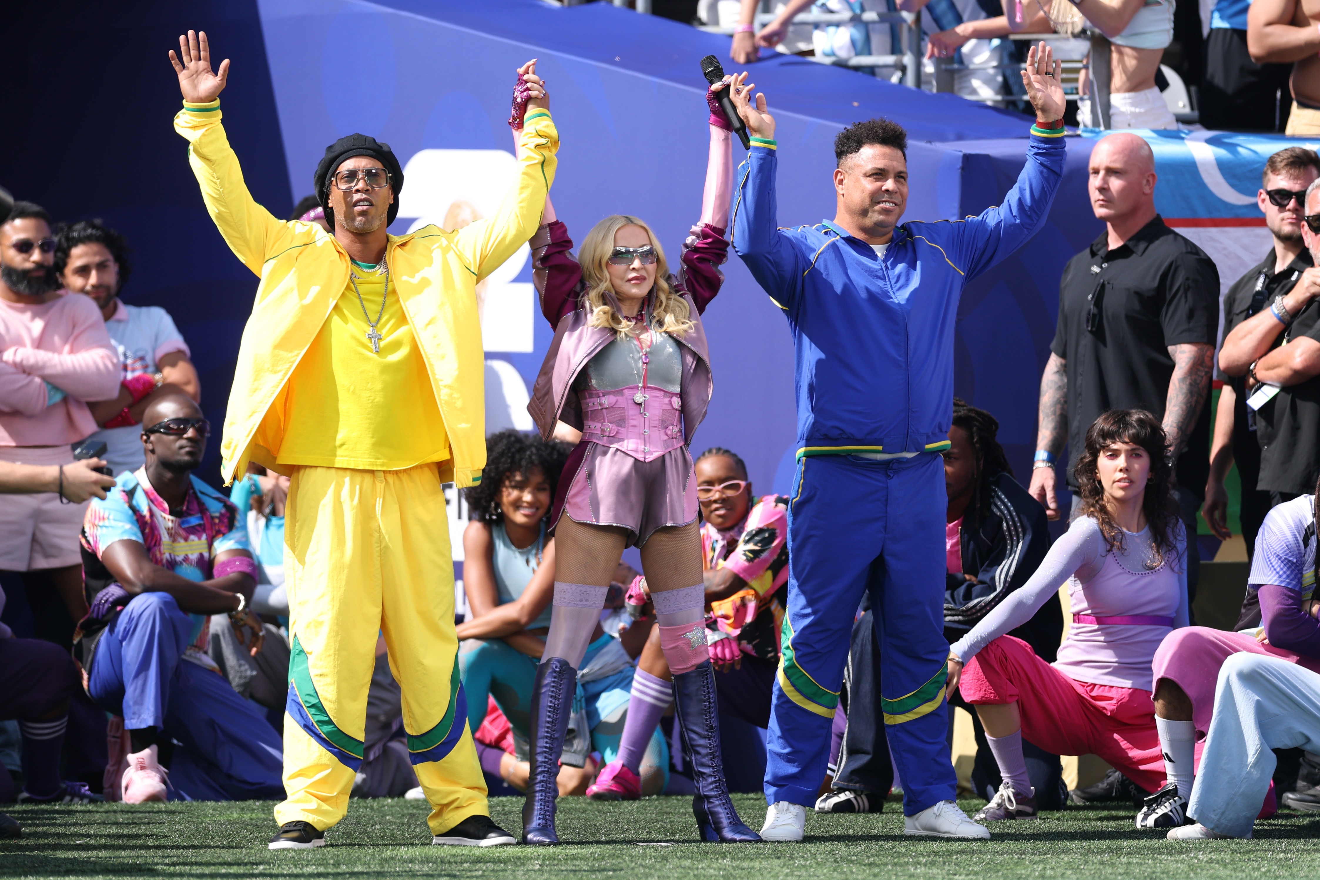 Ronaldinho Gaúcho, Madonna e Ronaldo Fenômeno no palco, braços erguidos, celebrando. Ronaldinho veste agasalho amarelo, Madonna um conjunto rosa com botas, e Ronaldo um agasalho azul. O público e seguranças estão ao fundo