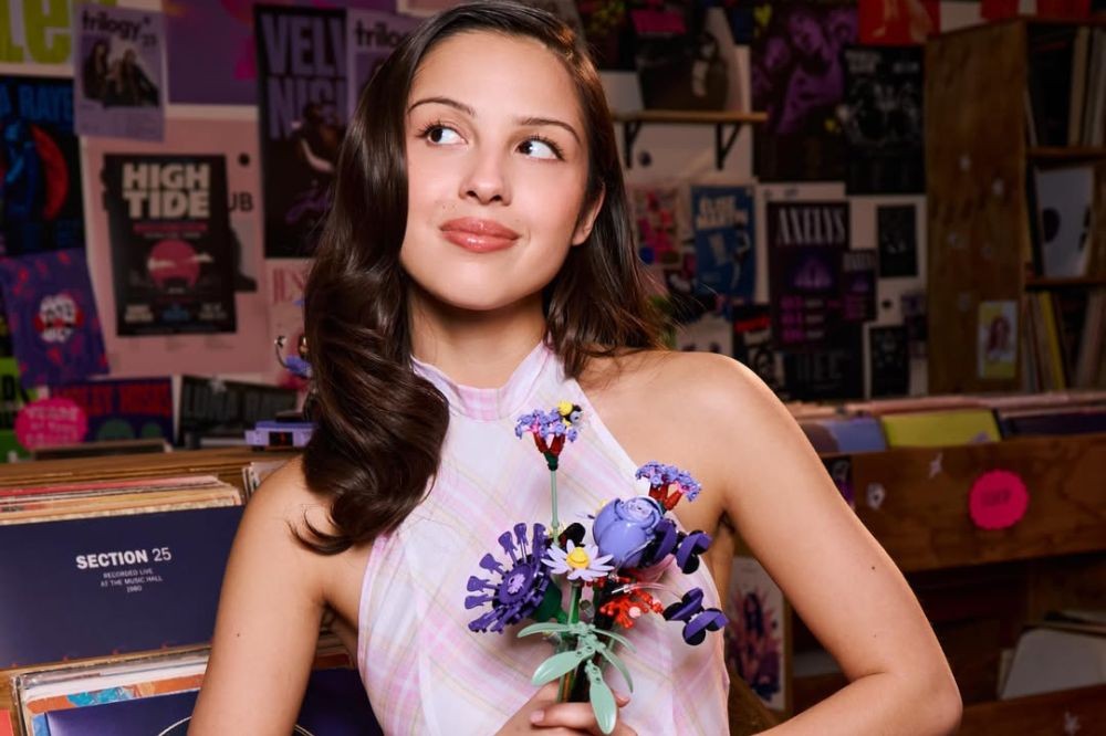 Olivia Rodrigo, jovem de cabelos castanhos e ondulados, sorri levemente e olha para cima, segurando um buquê de flores de Lego roxas e brancas. Ela veste um top xadrez rosa e branco, em uma loja de discos com pôsteres de bandas na parede