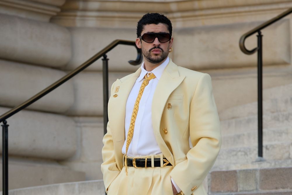 Bad Bunny posa em um terno creme com camisa branca, gravata dourada trançada, cinto preto e óculos escuros, em frente a uma escadaria de pedra com corrimão preto