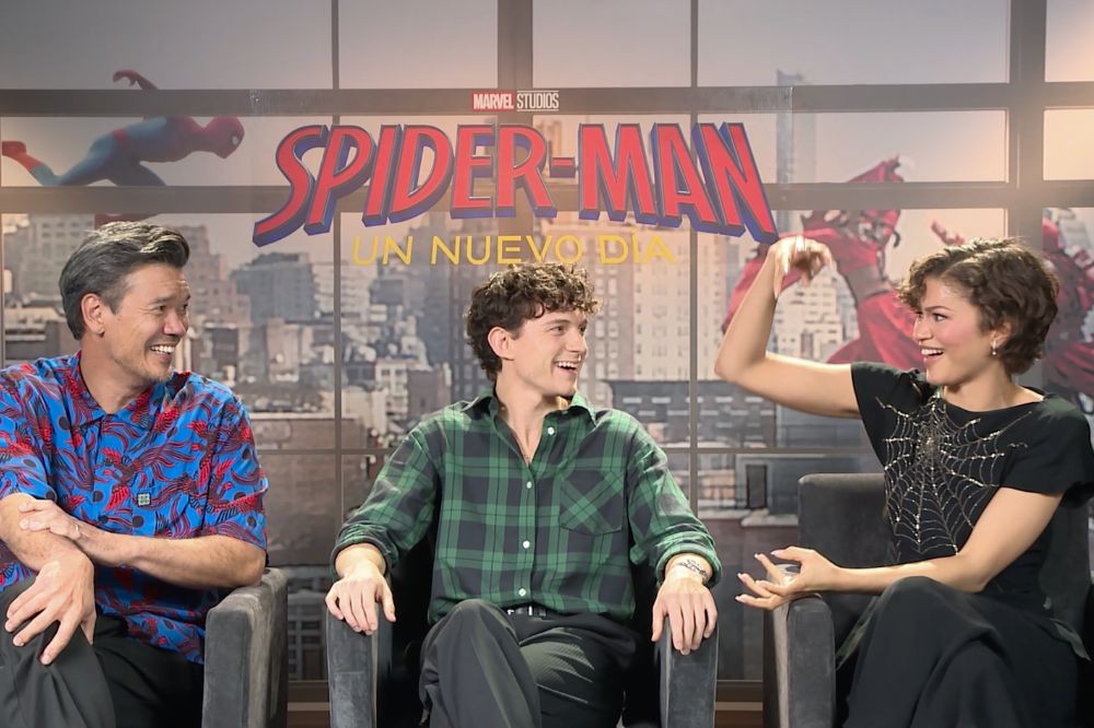 Três pessoas sorrindo e conversando sentadas em cadeiras escuras, com um banner de Spider-Man: Un Nuevo Día ao fundo. À esquerda, um homem de camisa azul e vermelha. No centro, um homem jovem de camisa xadrez verde. À direita, uma mulher de cabelo curto e blusa preta com teia de aranha, gesticulando com a mão direita levantada