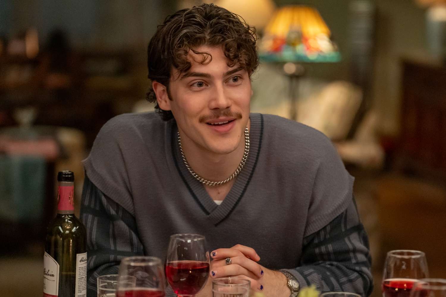 Homem jovem de cabelo castanho encaracolado e bigode, sorrindo e olhando para a direita. Ele veste um suéter cinza sobre uma camisa listrada e um colar prateado. Suas unhas estão pintadas de roxo escuro e ele usa um anel e um relógio. À frente, há taças de vinho e uma garrafa.