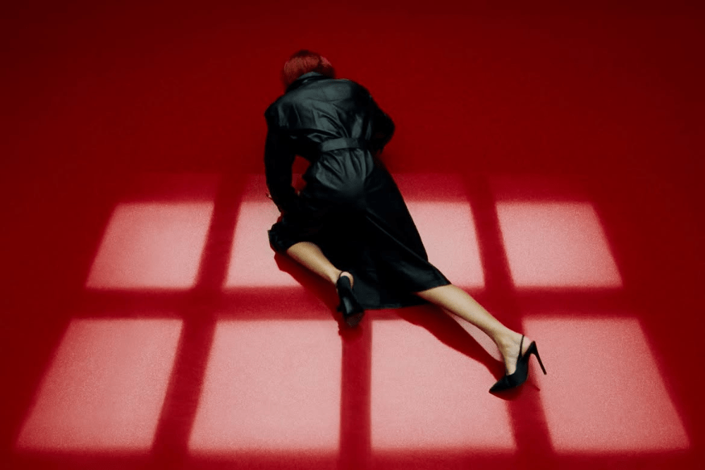 Mulher de cabelos ruivos e casaco preto, deitada de bruços no chão vermelho, com pernas esticadas e sapatos de salto alto. Luzes em formato de grade projetam sombras no chão.