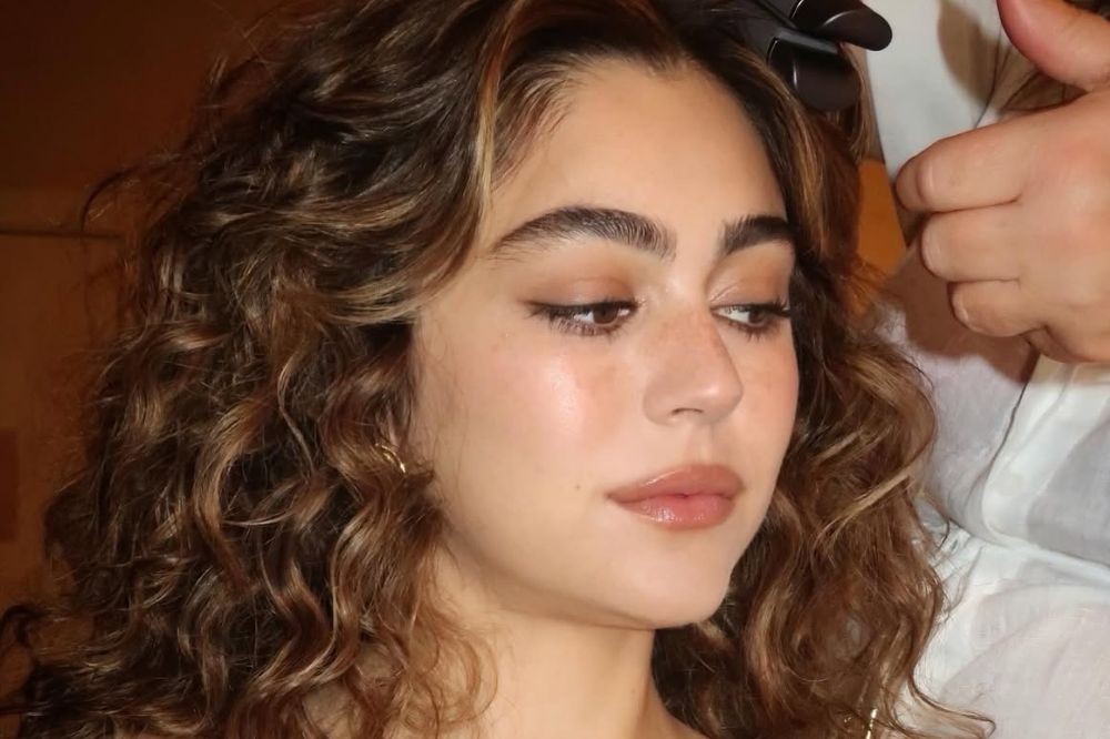 Mulher jovem com cabelos castanhos cacheados e mechas claras, maquiagem natural e delineador, olhando para baixo. Uma mão com luva preta segura seu cabelo no canto superior direito