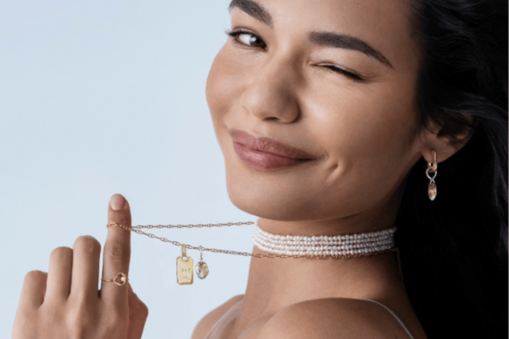 Mulher sorridente de pele morena e cabelo escuro, piscando o olho direito, usando um colar choker de pérolas, brincos e um anel com pedra, segurando um colar dourado com pingentes