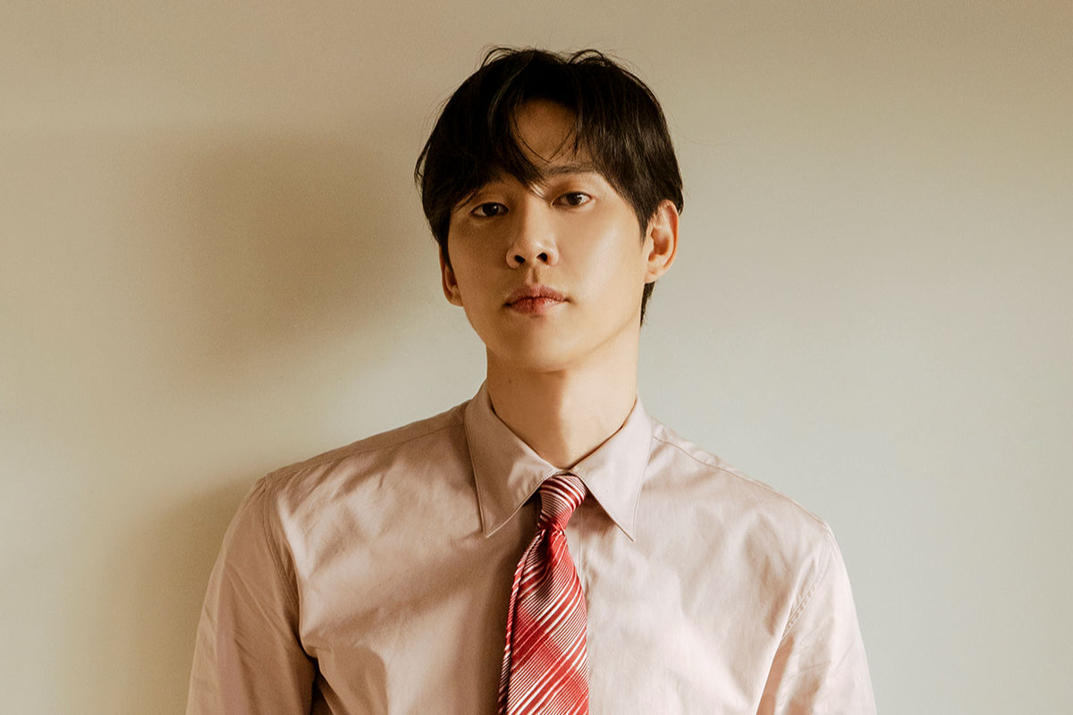 Homem asiático de cabelo escuro, camisa social rosa claro e gravata listrada vermelha e branca, olhando diretamente para frente com expressão séria