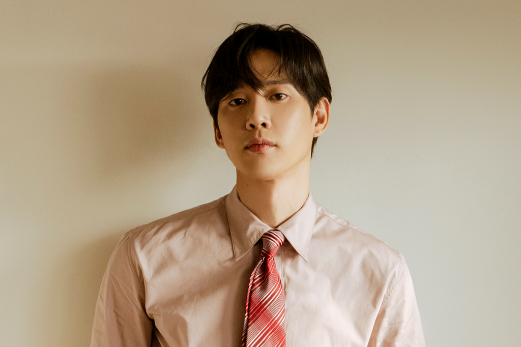 Homem asiático de cabelo escuro, camisa social rosa claro e gravata listrada vermelha e branca, olhando diretamente para frente com expressão séria