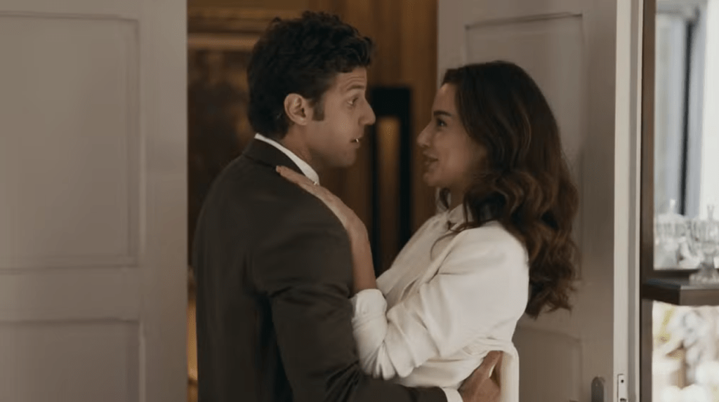 Um homem de terno escuro e uma mulher de blusa branca se abraçam, olhando um para o outro com sorrisos sutis, em um ambiente interno com portas e um espelho ao fundo