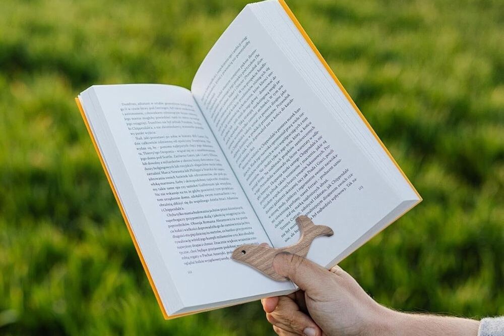 Mão segura um livro aberto com capa amarela e um marcador de página de madeira em formato de baleia, sobre fundo verde borrado de grama