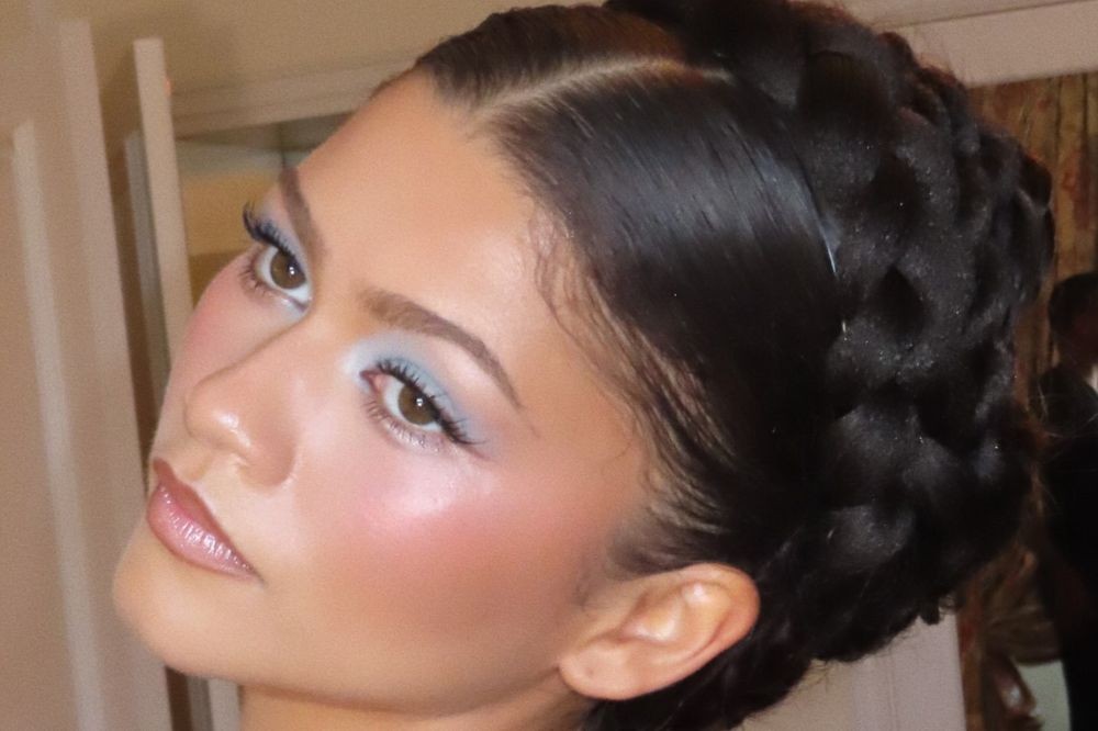 Mulher com pele bronzeada, maquiagem azul clara nos olhos e blush rosado, lábios com gloss. Cabelo escuro preso em uma trança grossa que contorna a cabeça, com risca lateral e baby hair. Ela olha para cima, com a cabeça levemente inclinada para a esquerda