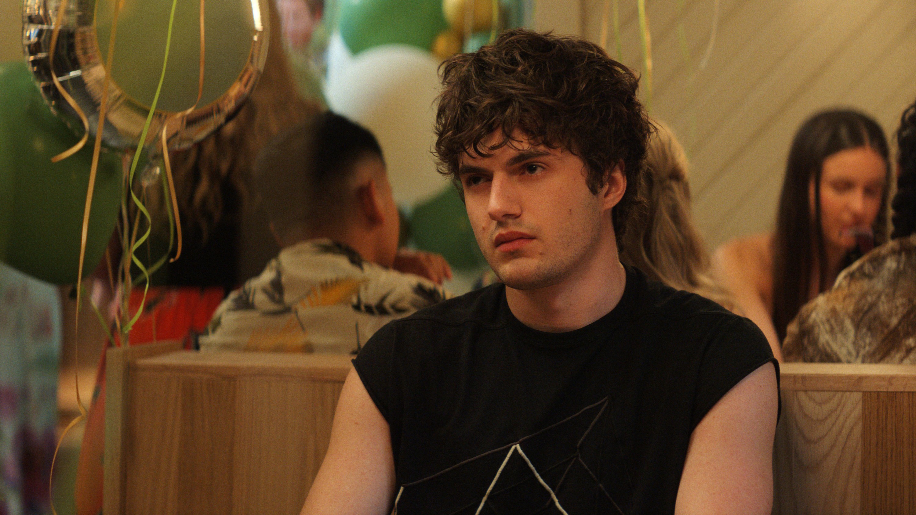 Jovem de cabelo cacheado escuro e camiseta preta com estampa geométrica branca, sentado em um ambiente festivo com balões verdes e dourados, olhando para a direita com expressão séria