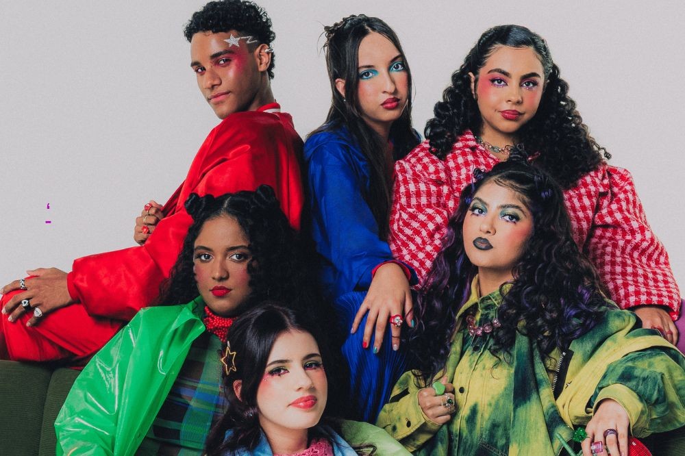 Seis jovens, três em pé e três sentados, posam com maquiagens coloridas e roupas vibrantes. O homem de vermelho tem uma estrela prateada no rosto. As mulheres usam blusas azul, xadrez vermelho, verde brilhante, xadrez verde e tie-dye verde e amarelo. Todos olham para a câmera com expressões sérias
