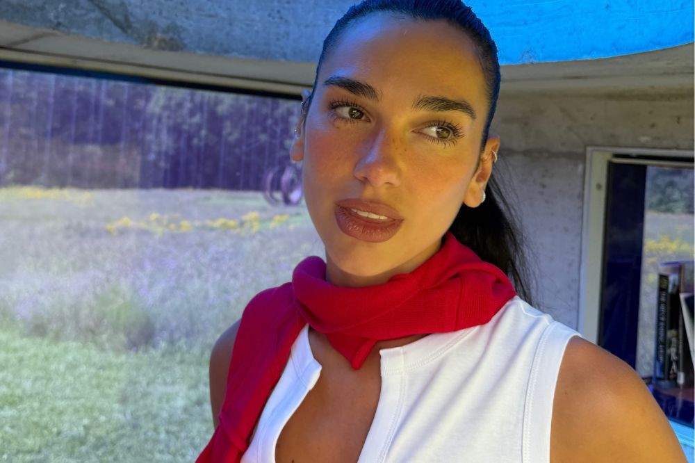 Dua Lipa com cabelo escuro preso, maquiagem leve e sardas, usando regata branca e suéter vermelho amarrado no pescoço, olhando para a direita com um fundo de paisagem verde e azul