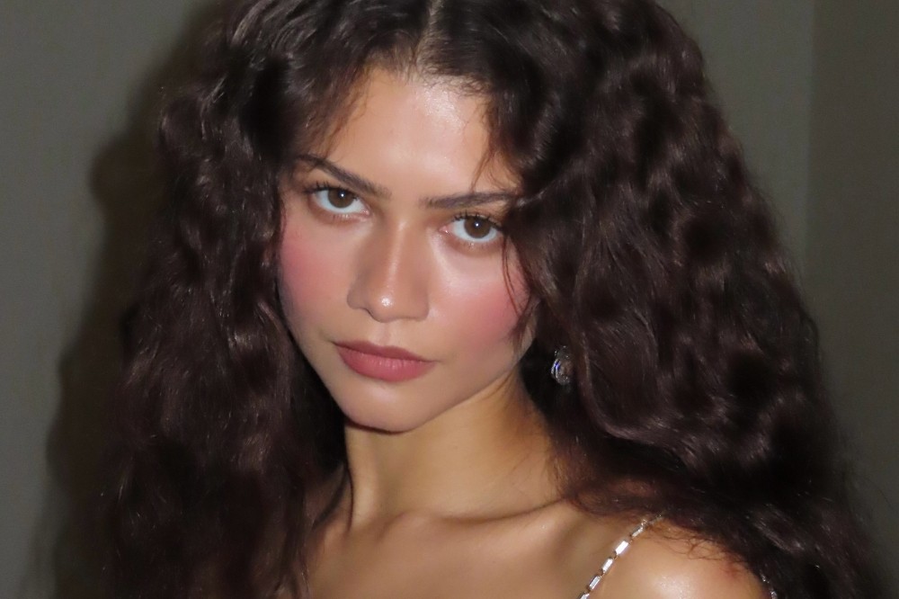 Rosto da atriz Zendaya, com cabelos castanhos escuros e ondulados, maquiagem rosa nas bochechas e lábios, e um brinco brilhante na orelha direita. Ela olha diretamente para a câmera com uma expressão séria