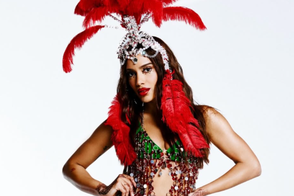 Mulher de pele morena, cabelo castanho escuro, usa um cocar de penas vermelhas e pedrarias prateadas, e um top verde com franjas de miçangas vermelhas e prateadas. Ela tem maquiagem brilhante nos olhos e batom vermelho, com as mãos na cintura, em fundo branco