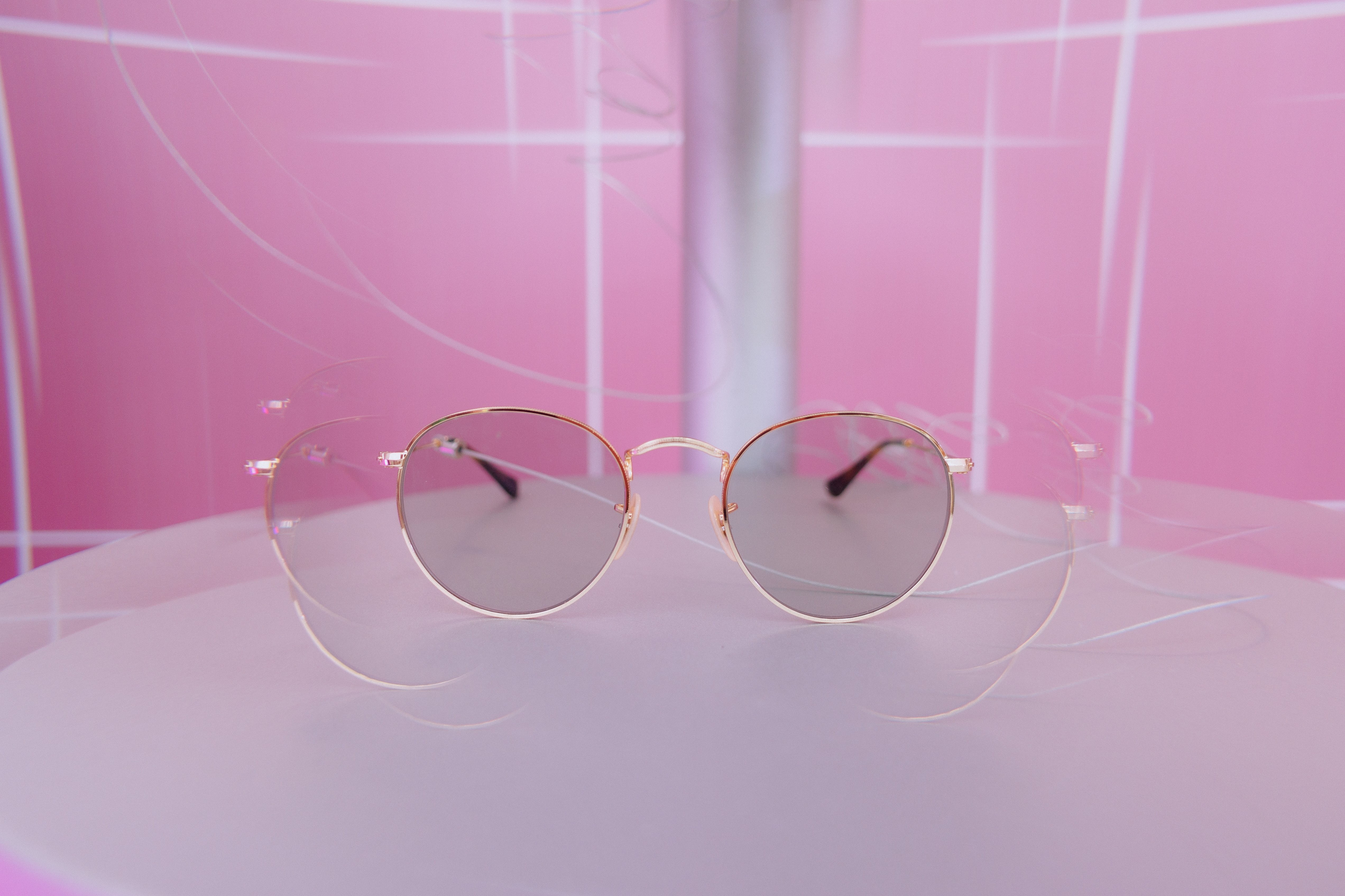 Óculos de sol com armação dourada e lentes espelhadas cinza, apoiados em uma superfície lilás clara, com fundo rosa e linhas brancas