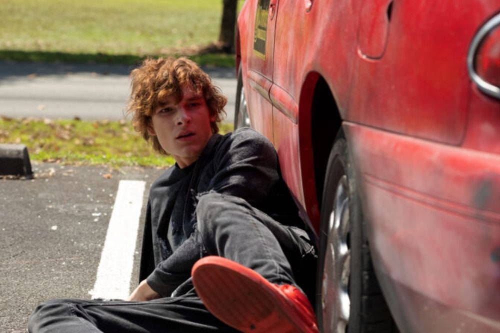 Jovem de cabelo ruivo encaracolado, vestindo jaqueta e calça escuras, tênis vermelho, sentado no chão de um estacionamento, encostado em um carro vermelho, olhando para o lado com expressão preocupada