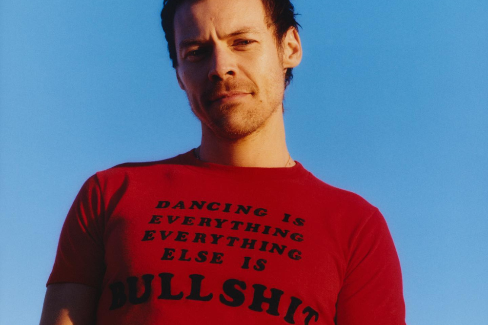 Homem de cabelo escuro e barba rala, vestindo camiseta vermelha com a frase DANCING IS EVERYTHING EVERYTHING ELSE IS BULLSHIT em preto, olhando para frente com expressão séria, contra um céu azul claro