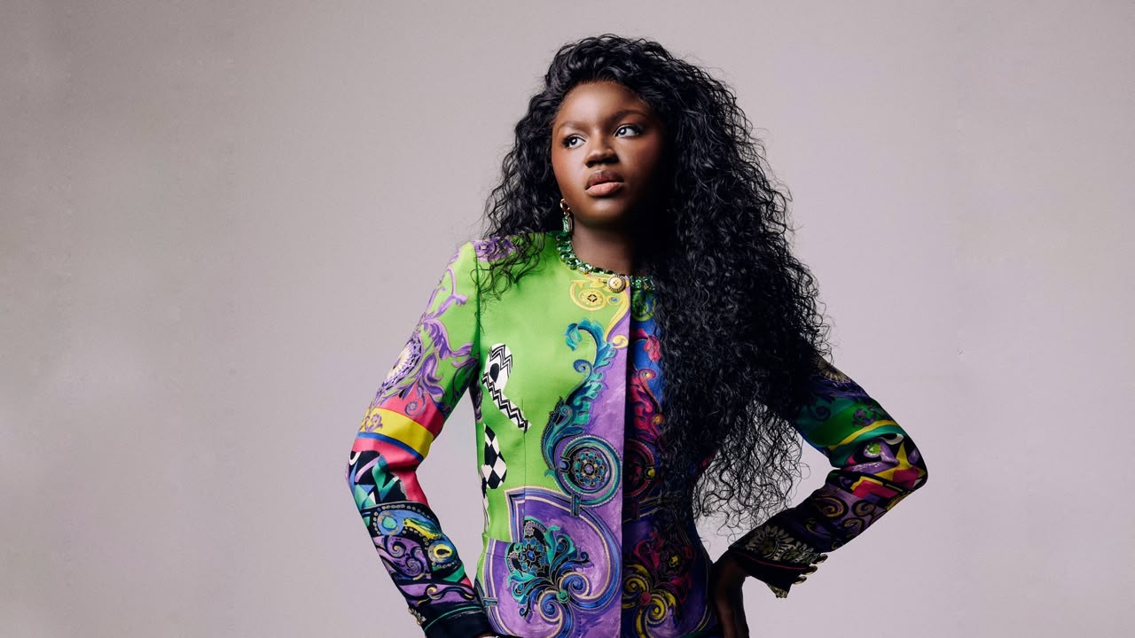 Mulher negra de cabelos longos e cacheados, vestindo um blazer vibrante com estampas barrocas em tons de verde, roxo, rosa e amarelo, posa com as mãos na cintura e olhar sério para o lado direito, contra um fundo cinza claro
