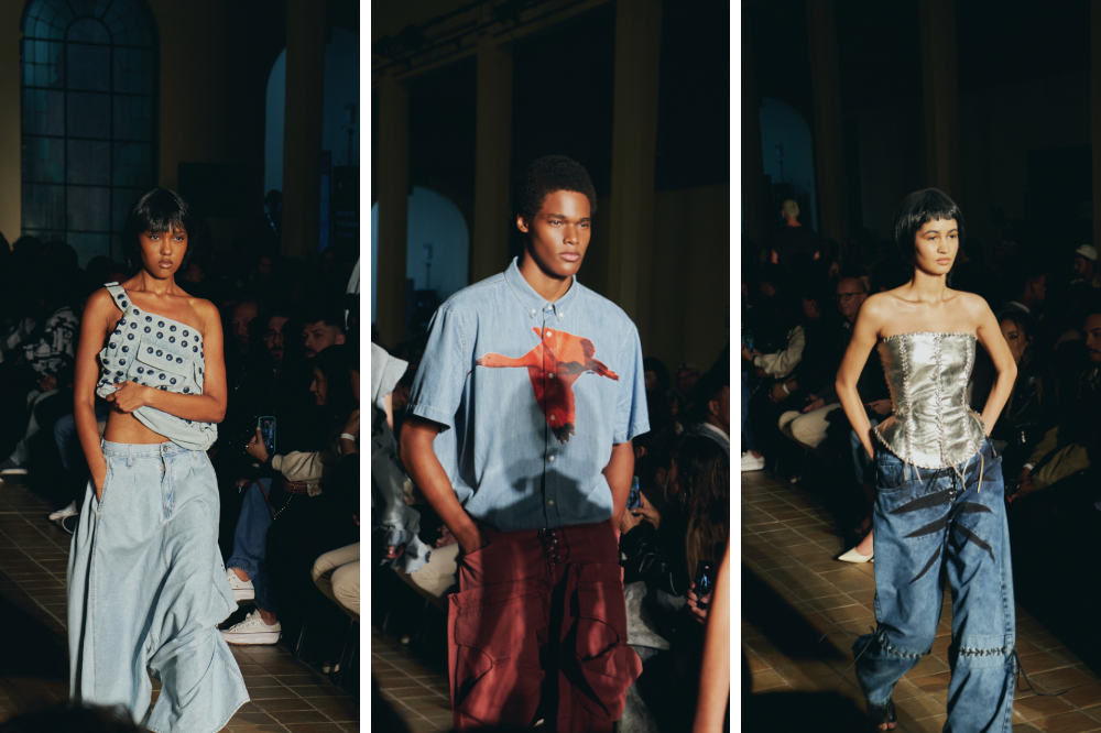 Três modelos desfilam em passarela escura. Uma mulher veste top de um ombro só com bolinhas e calça jeans larga. Um homem usa camisa azul clara com estampa laranja e calça marrom. Outra mulher veste corpete prateado e calça jeans com detalhes pretos e amarrações