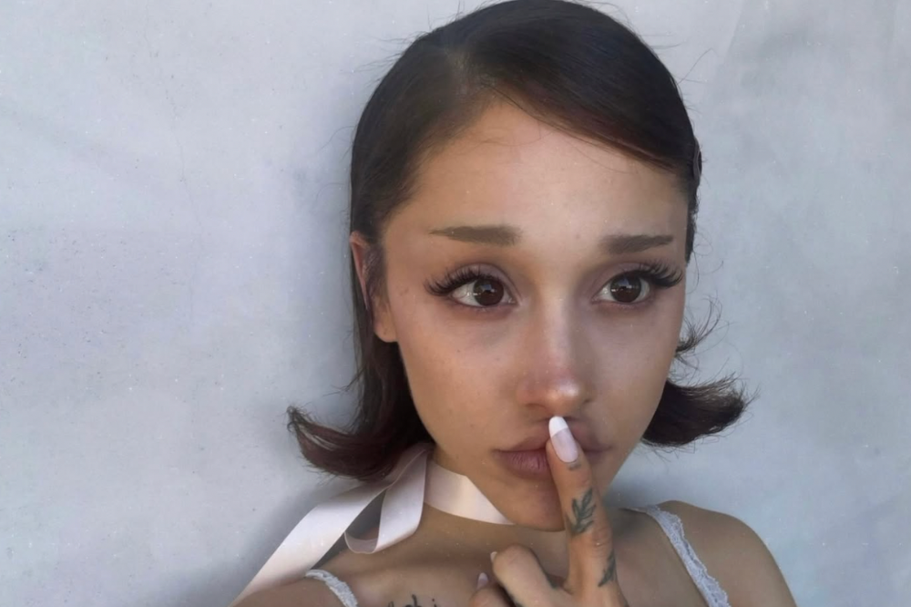 Cantora Ariana Grande com cabelo castanho escuro e maquiagem leve, usando um top branco de renda e fita rosa no pescoço, com o dedo indicador na boca, fazendo sinal de silêncio