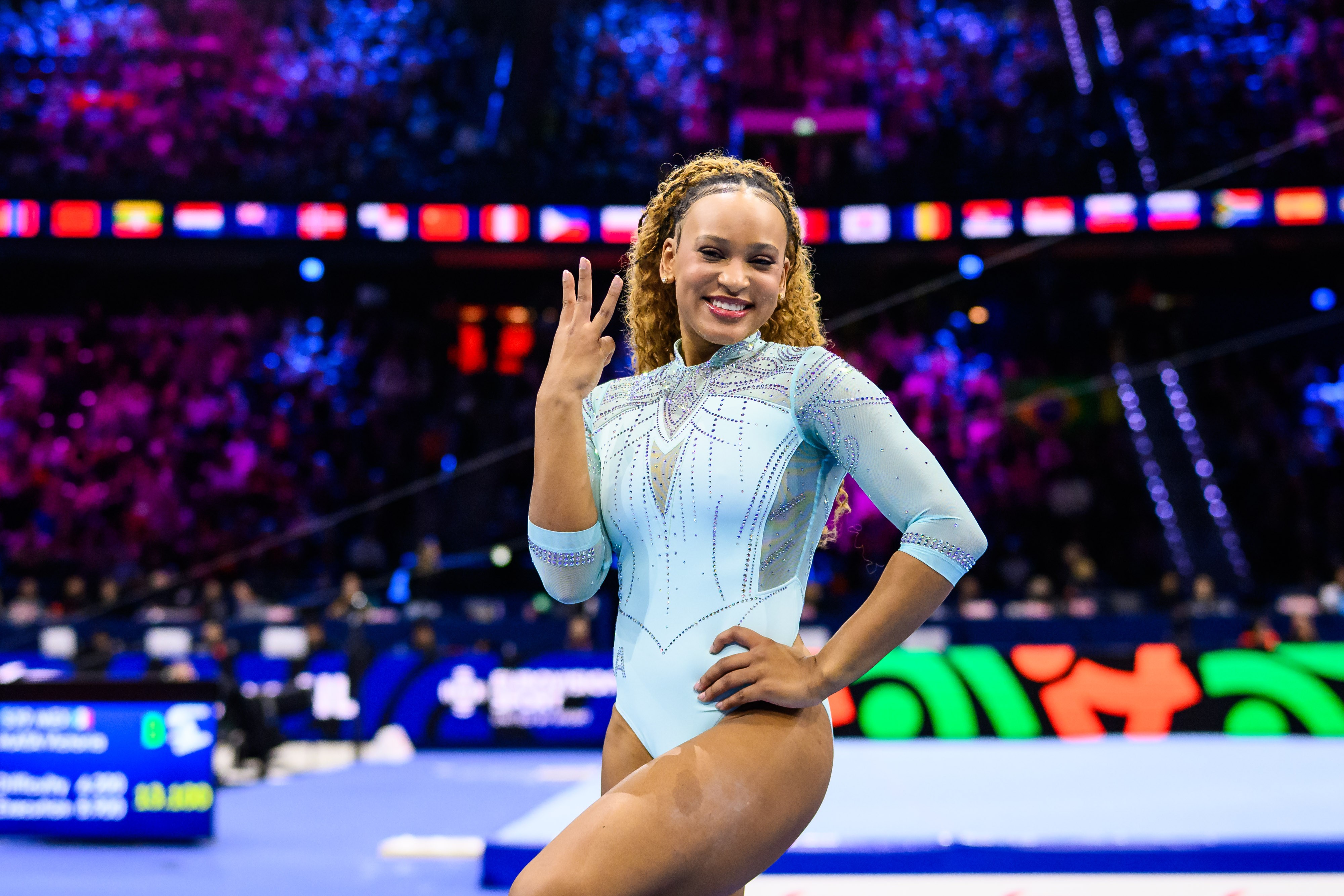 Ginasta Rebeca Andrade sorrindo, com cabelo cacheado loiro, vestindo um collant azul-claro brilhante, fazendo um sinal de três com a mão direita, com a mão esquerda na cintura, em uma arena esportiva com público e luzes coloridas ao fundo